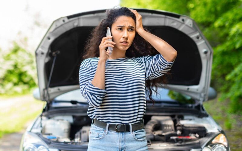a young woman talking on the phone with a car’s open hood in the background